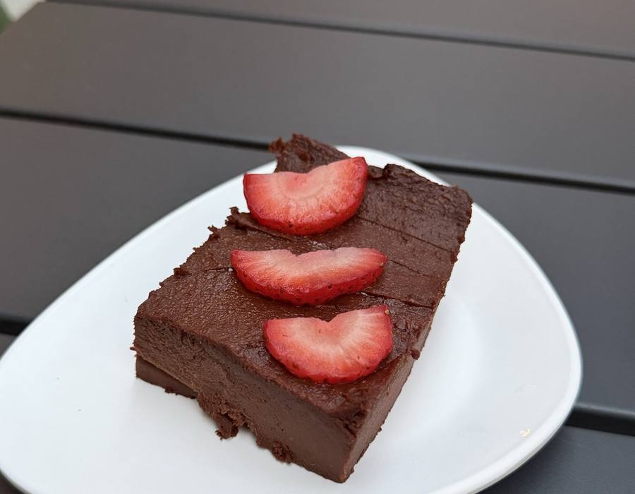 A thick square of dark chocolate brownie on a white plate, topped with three fanned slices of fresh strawberry