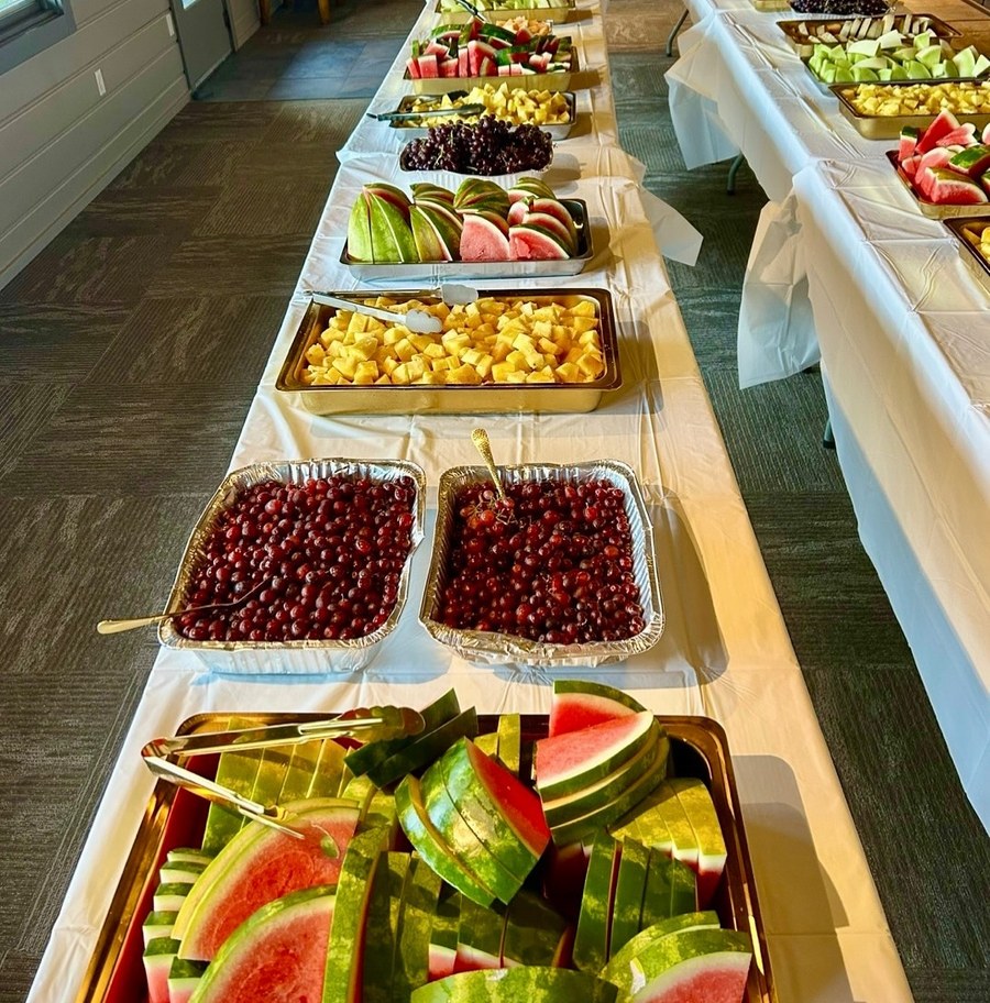Two long banquet tables lined end to end with trays of fruit -- watermelon, cherries, pineapple, grapes, honeydew, apples and melon