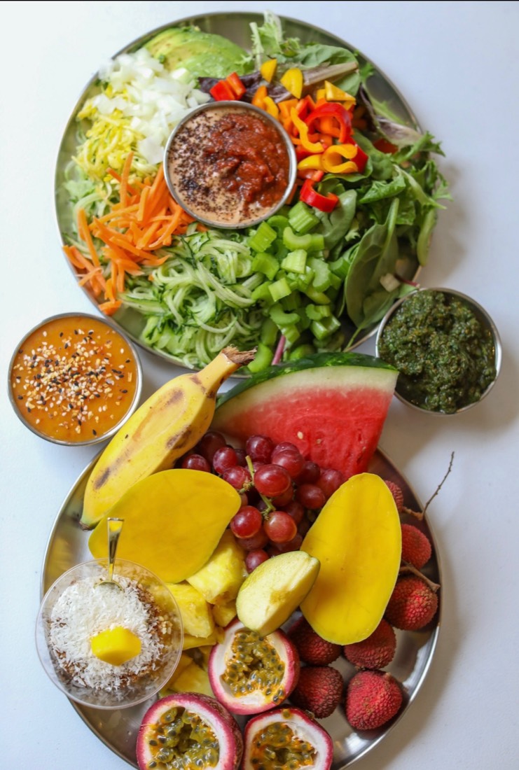 Two steel platters side by side: the salad bar with mixed greens, avocado, shredded carrot, courgette noodles, celery, sweet peppers and three dressings; and the fruit bar with mango, lychee, passion fruit, watermelon, pineapple, grapes, banana and shredded coconut